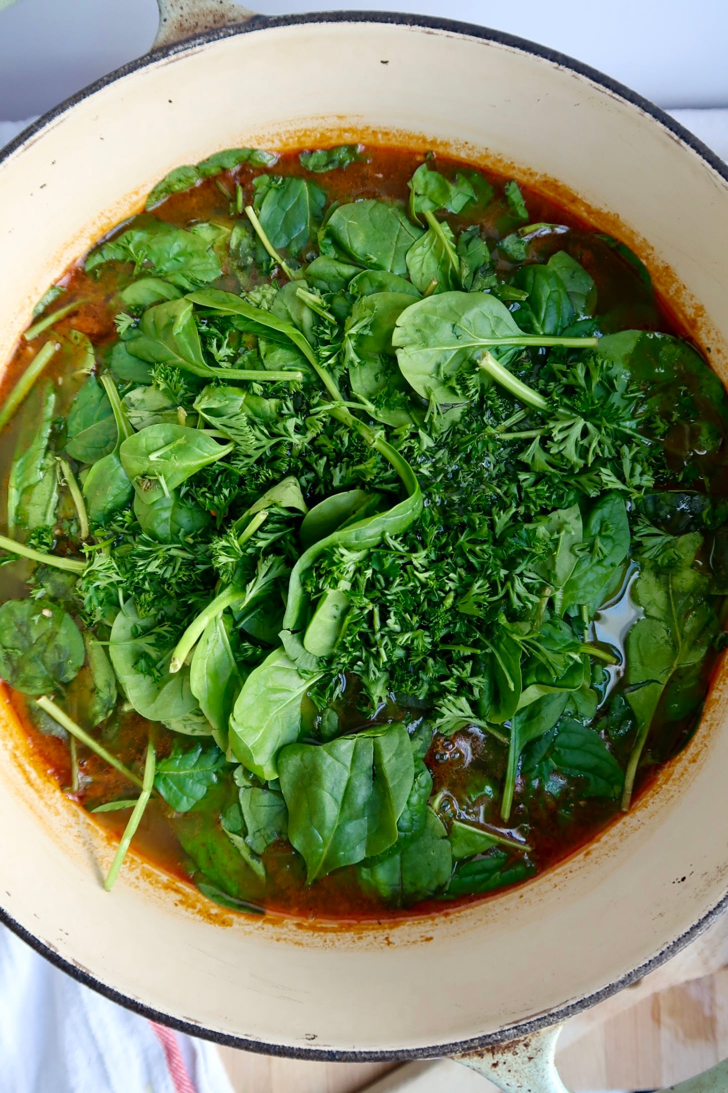 A pot of soup with fresh spinach leaves and chopped parsley on top, sitting in a reddish broth, ready to be stirred and cooked.