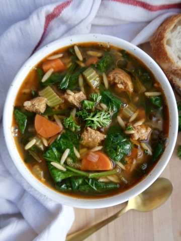 A bowl of vegetable and chicken soup with carrots, celery, greens, orzo pasta, and herbs, placed on a wooden surface next to a slice of bread and a gold spoon, with a white and red cloth nearby.