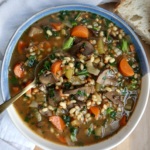 A bowl of vegetable and lentil soup with carrots, mushrooms, greens, and a spoon, next to a slice of rustic bread on a wooden surface with a white cloth.