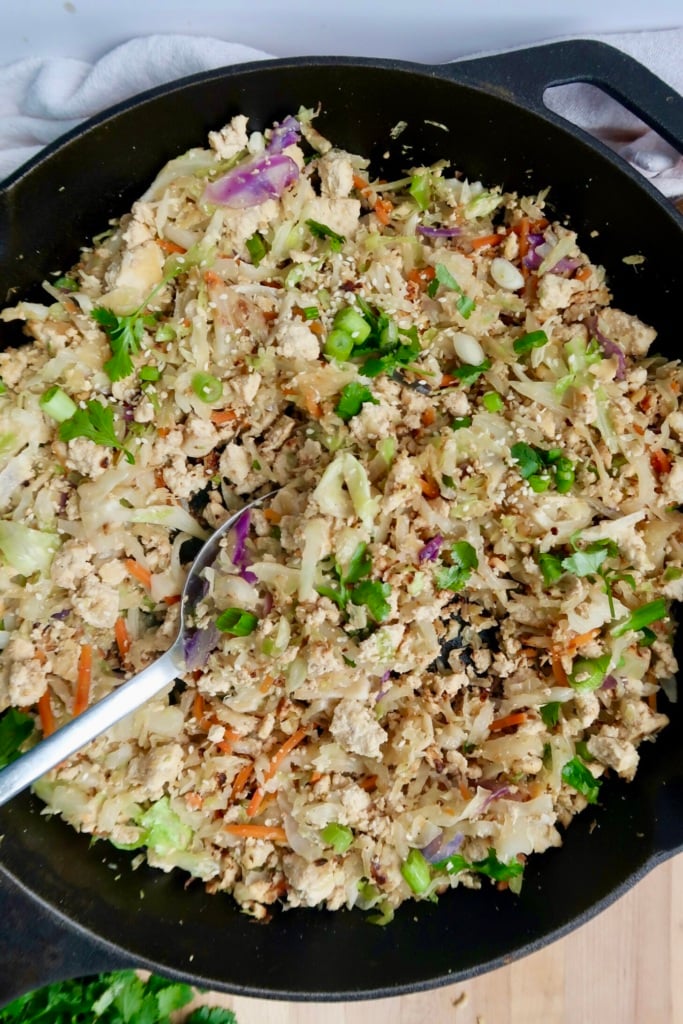 A skillet filled with a cooked mixture of ground meat, shredded cabbage, carrots, green onions, and herbs, with a spoon resting inside the pan.