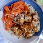 Bowl of rice with baked tofu with a teriyaki sauce and ribboned carrots with sesame seeds on top.