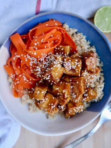 Bowl of rice with baked tofu with a teriyaki sauce and ribboned carrots with sesame seeds on top.