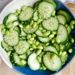 White and blue bowl with sliced cucumber and edamame mixed together with sesame seeds on top.