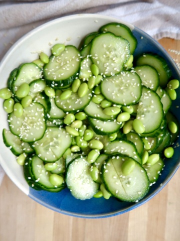 White and blue bowl with sliced cucumber and edamame mixed together with sesame seeds on top.