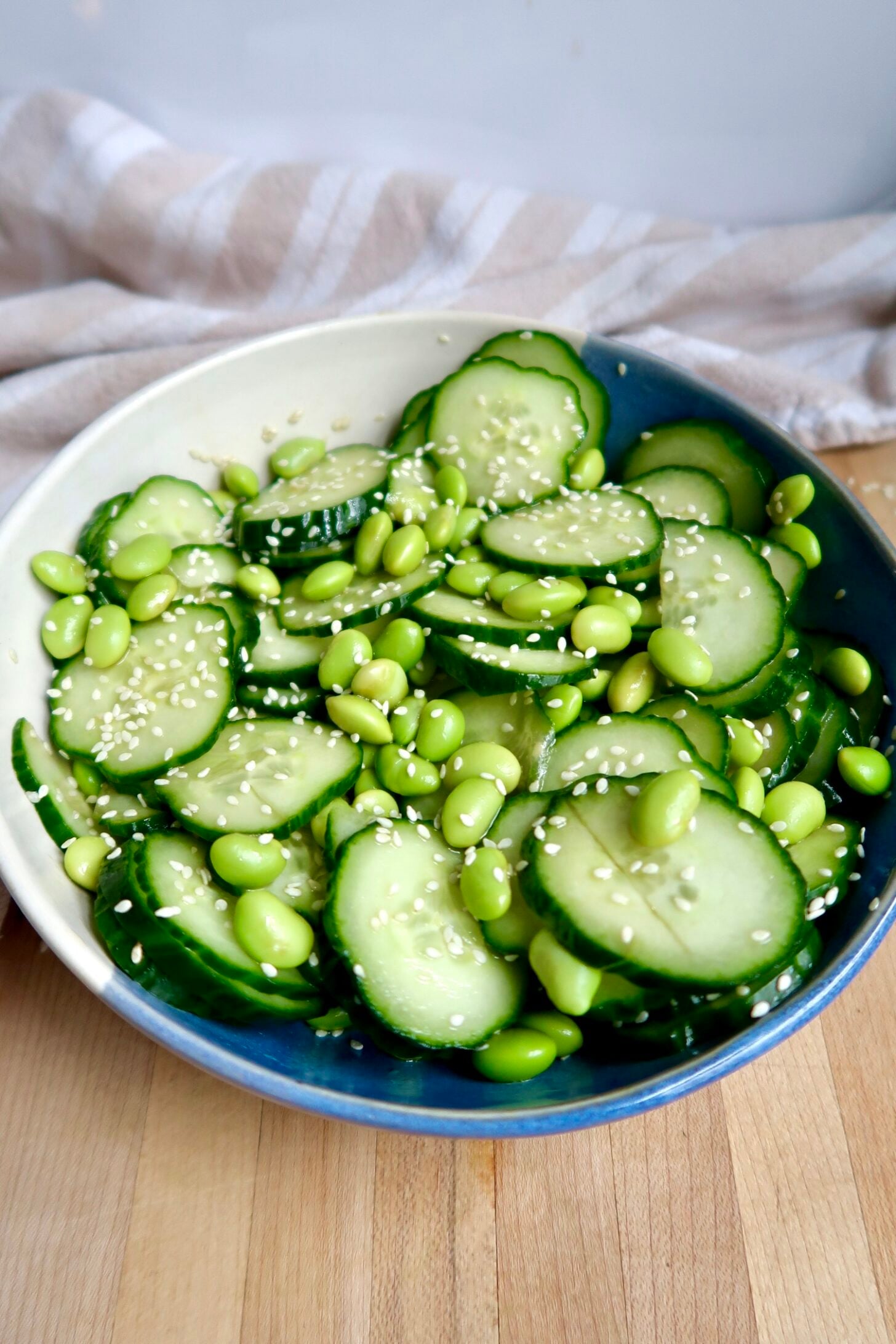 Side view of White and blue bowl with sliced cucumber and edamame mixed together with sesame seeds on top.