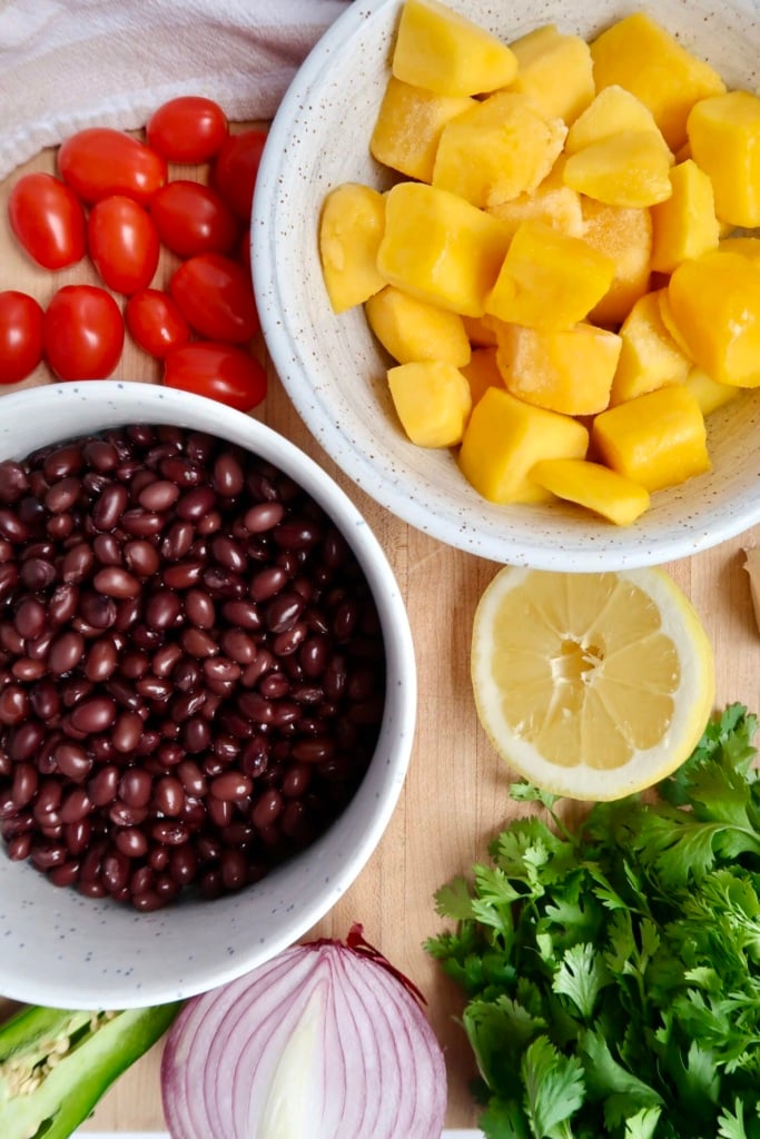 Cutting board with a bowl of mango, black beans, cherry tomatoes, red onion, jalapeno, cilantro and garlic laid out as ingredients.