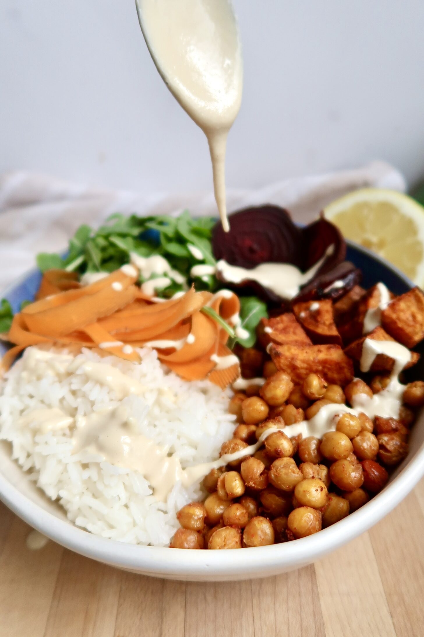 Bowl with rice, roasted chickpeas, beets, shaved carrot and arugula with a creamy dressing being drizzled on top.