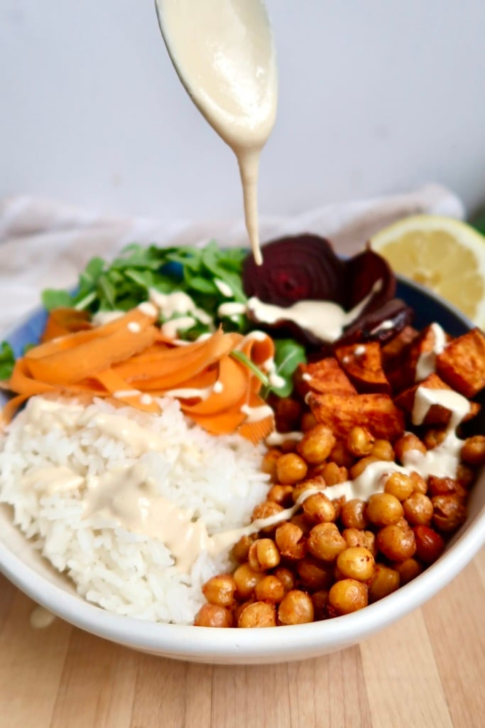 Bowl with roasted chickpeas, beets, rice, carrot, arugula and a creamy sauce being poured on top.