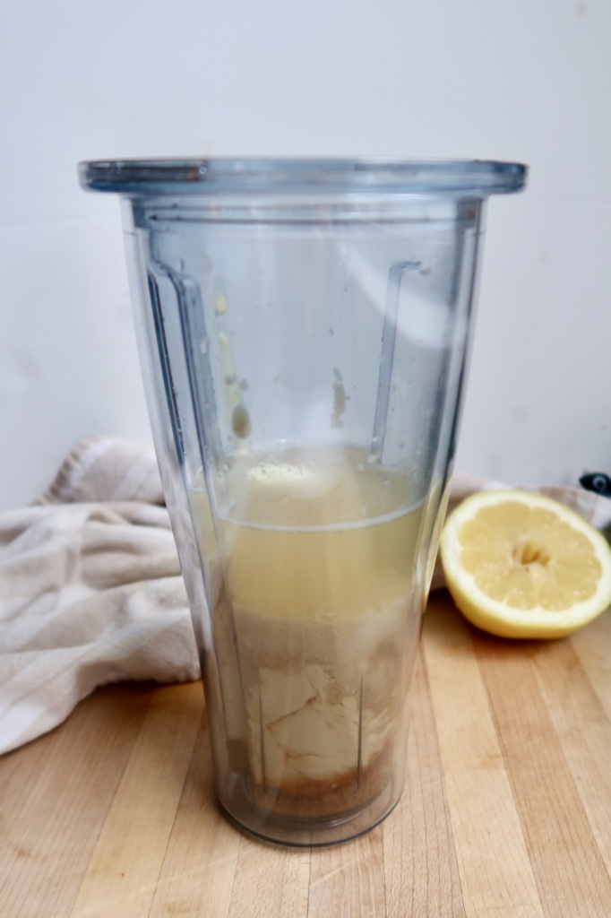 Blender container with tahini, lemon juice, garlic layered and a lemon off to the side.