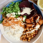 Bowl with rice, roasted chickpeas, roasted sweet potato, beets, arugula and carrot with a creamy sauce on top.