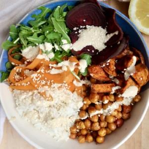 Bowl with rice, roasted chickpeas, roasted sweet potato, beets, arugula and carrot with a creamy sauce on top.