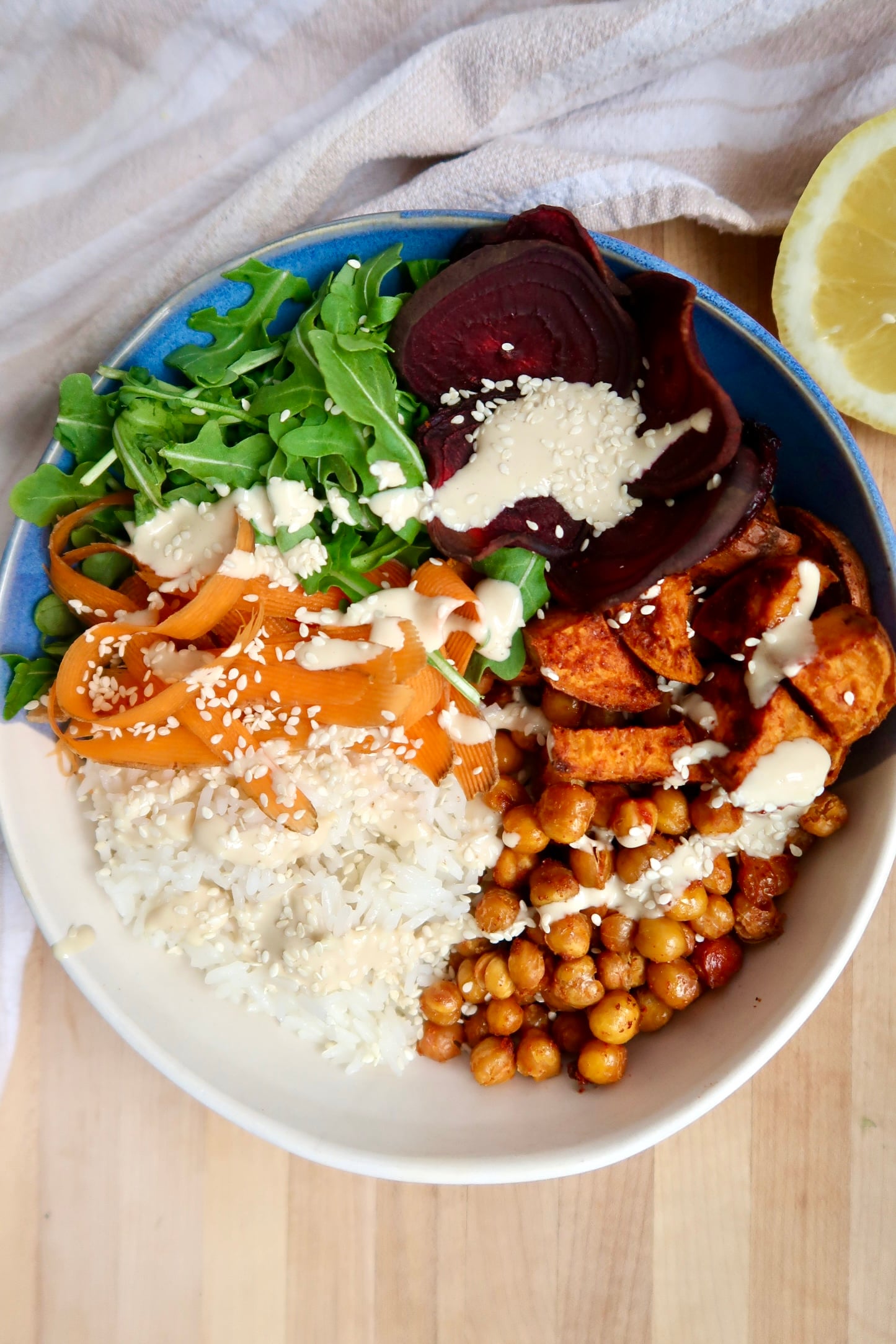 Bowl with rice, roasted chickpeas, roasted sweet potato, beets, arugula and carrot with a creamy sauce on top.