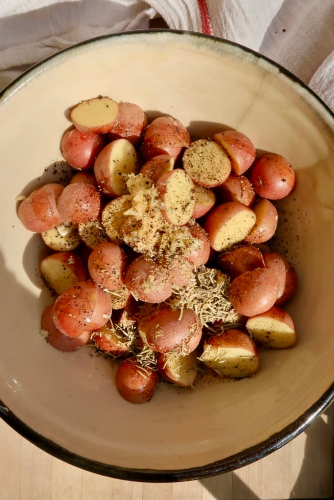 Halved red potatoes in a bowl with seasonings like fresh garlic and rosemary on them.