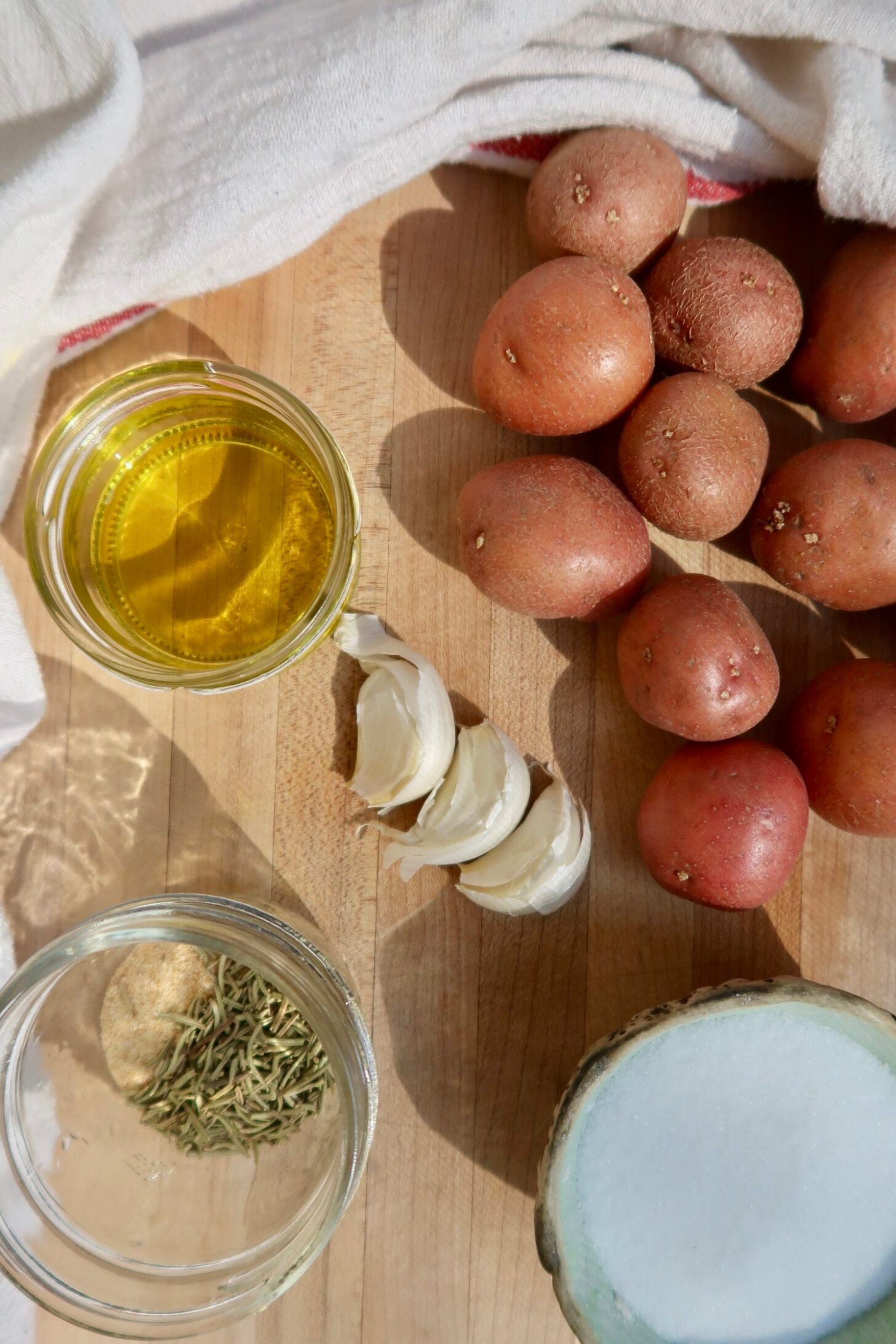 Olive oil, red potatoes, garlic, rosemary and salt on a cutting board.