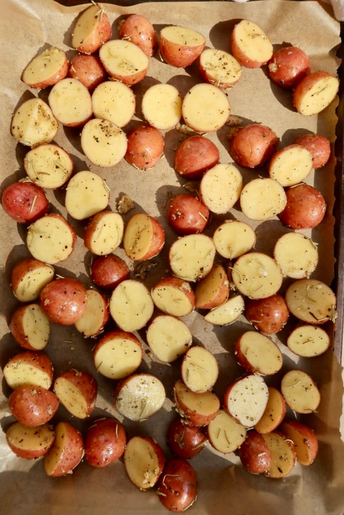 Halved red potatoes on a sheet pan with parchment paper and rosemary on them.