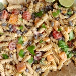 Creamy pasta salad with red bell peppers, cherry tomatoes, red onion, cilantro and a lime off to the side with salad tongs.