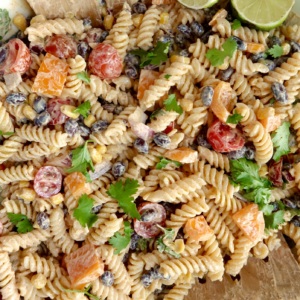 Creamy pasta salad with red bell peppers, cherry tomatoes, red onion, cilantro and a lime off to the side with salad tongs.