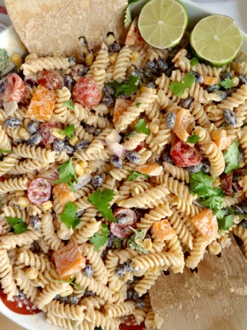 Creamy pasta salad with red bell peppers, cherry tomatoes, red onion, cilantro and a lime off to the side with salad tongs.