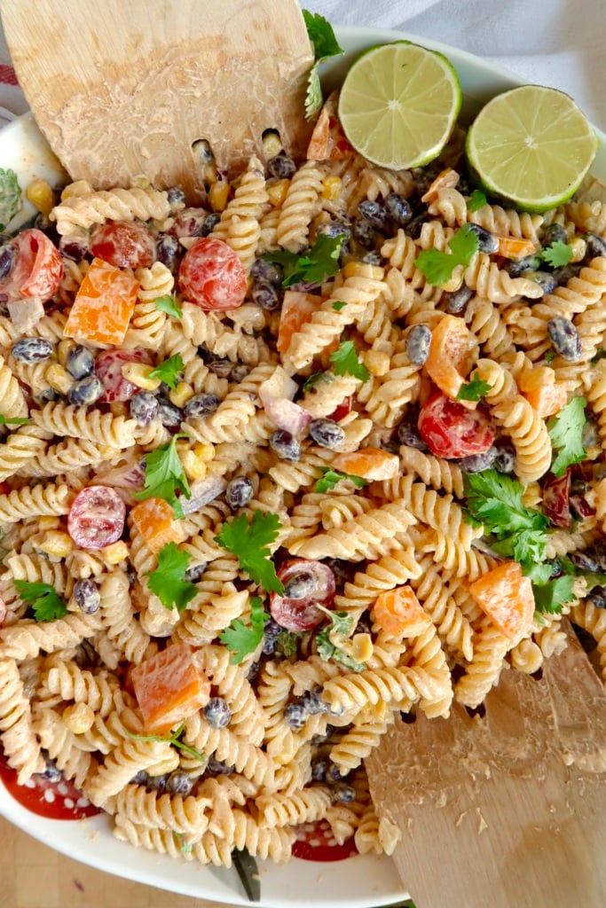 Creamy pasta salad with red bell peppers, cherry tomatoes, red onion, cilantro and a lime off to the side with salad tongs.