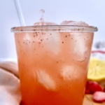 A glass of pink iced drink with visible ice cubes and black specks, a straw, and a pale napkin on a wooden surface, with a lemon slice and strawberries in the background. Liquid is being poured into the glass.