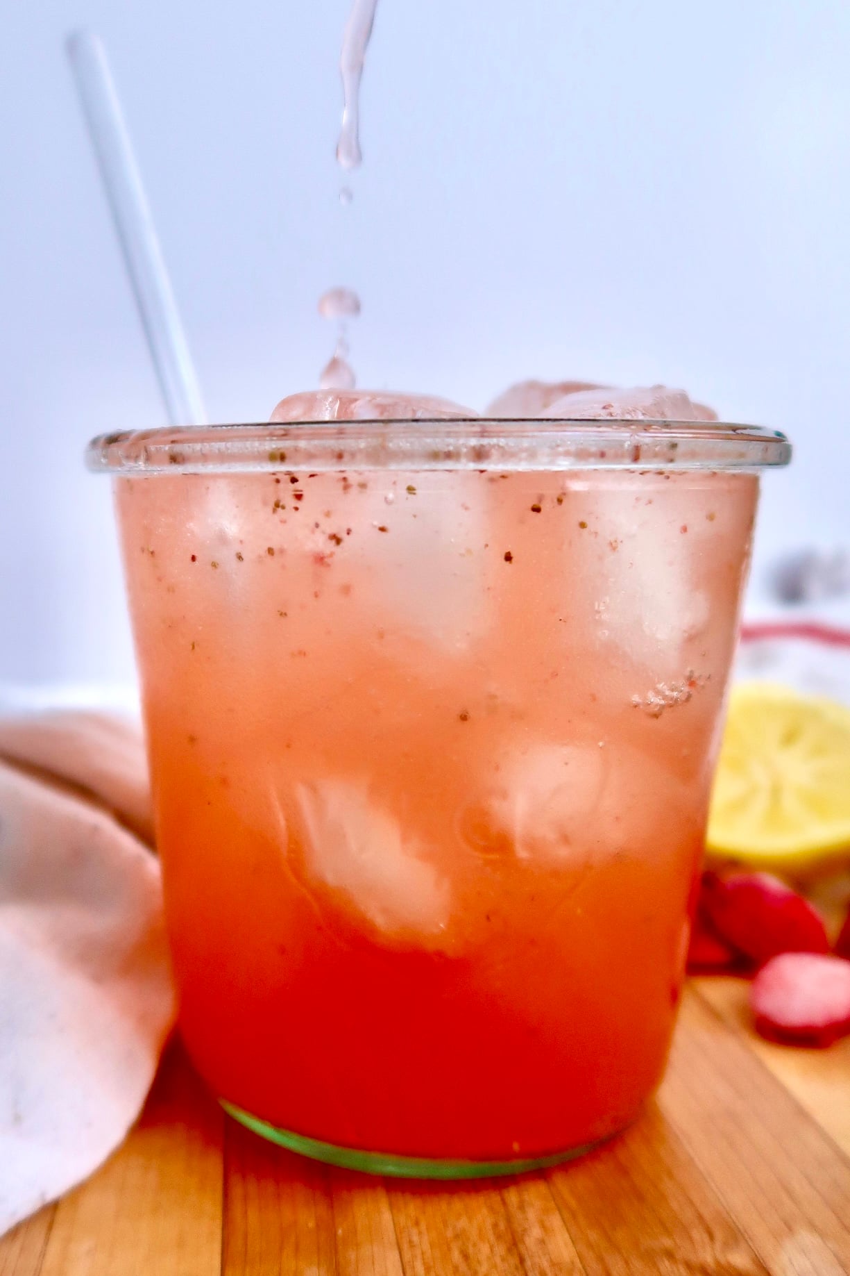 A glass of pink iced drink with visible ice cubes and black specks, a straw, and a pale napkin on a wooden surface, with a lemon slice and strawberries in the background. Liquid is being poured into the glass.