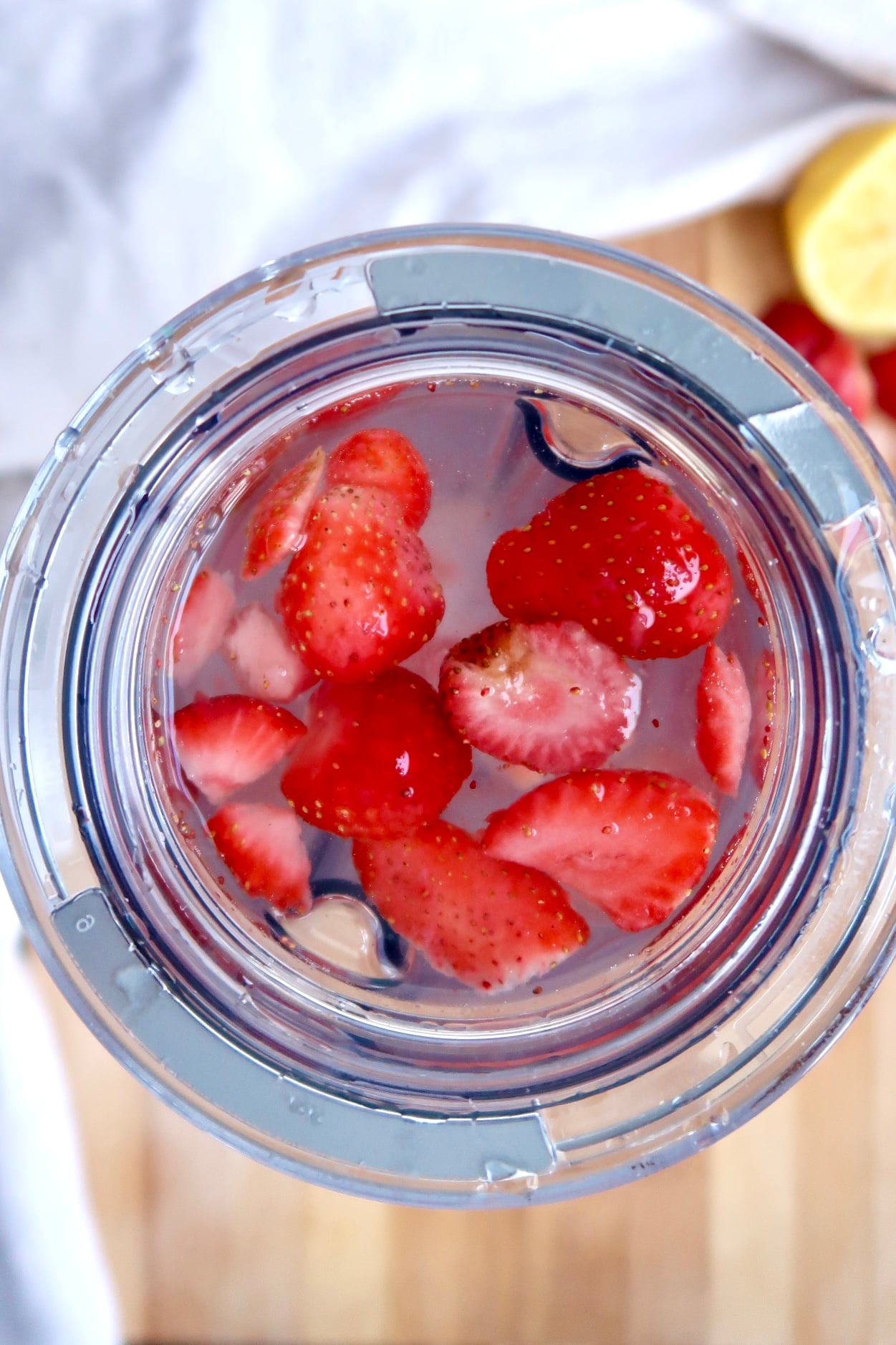 Top-down view of a clear container filled with water and sliced strawberries, with part of a lemon and whole strawberries visible on a wooden surface in the background.