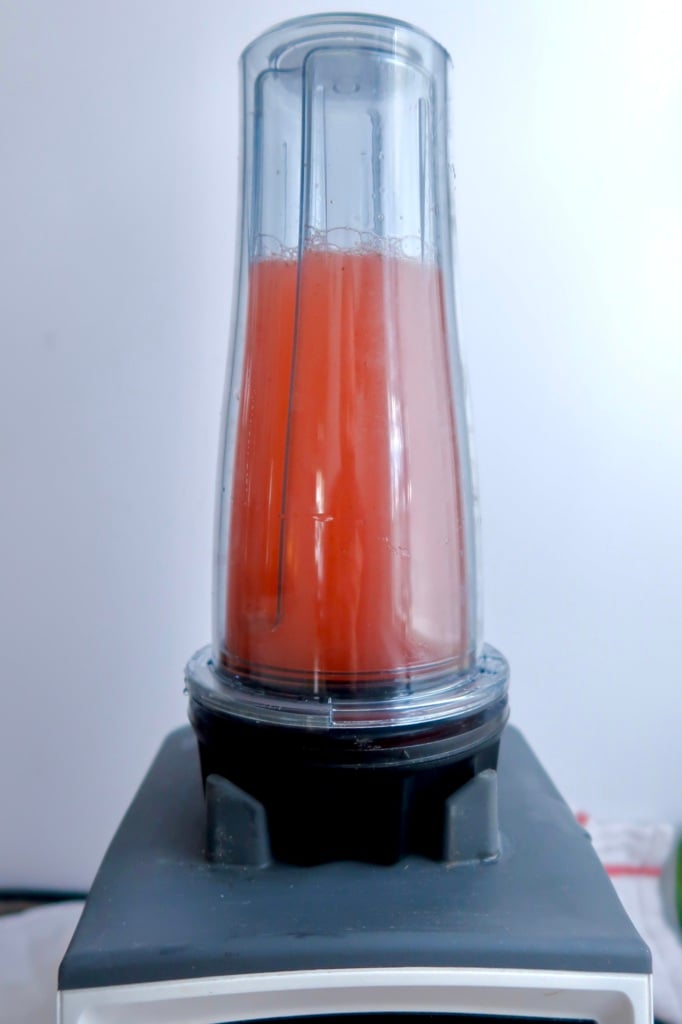 A close-up of a blender mixing a red liquid, juice or smoothie, inside a clear blending cup placed upside down on the blender base against a plain background.