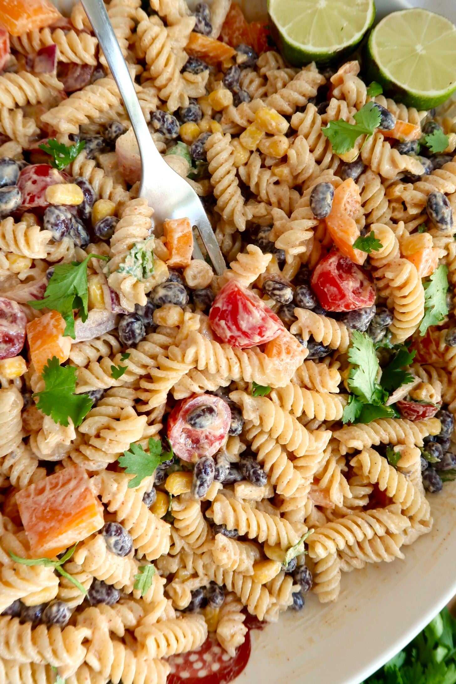 Creamy pasta salad with red bell peppers, cherry tomatoes, red onion, cilantro and a lime off to the side and a fork piercing some pasta.