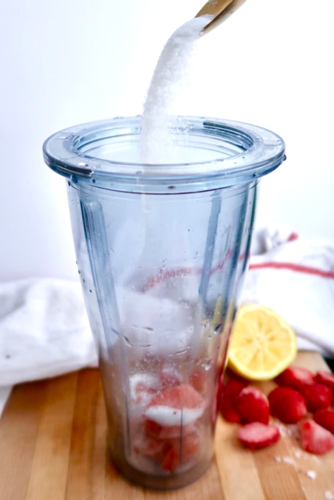 A clear blender cup with strawberries inside sits on a wooden surface. A spoon is pouring sugar into the cup. Sliced strawberries and half a lemon are on the table nearby. A white towel is in the background.