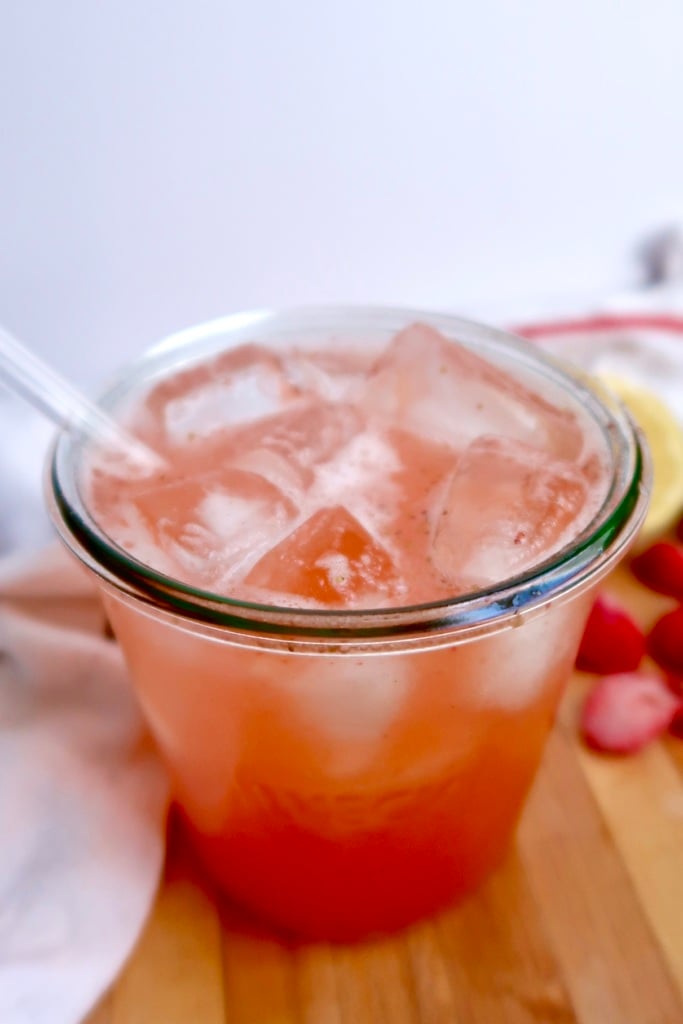 A glass filled with a pink iced drink and a clear straw sits on a wooden surface, with sliced strawberries and a lemon in the blurred background.