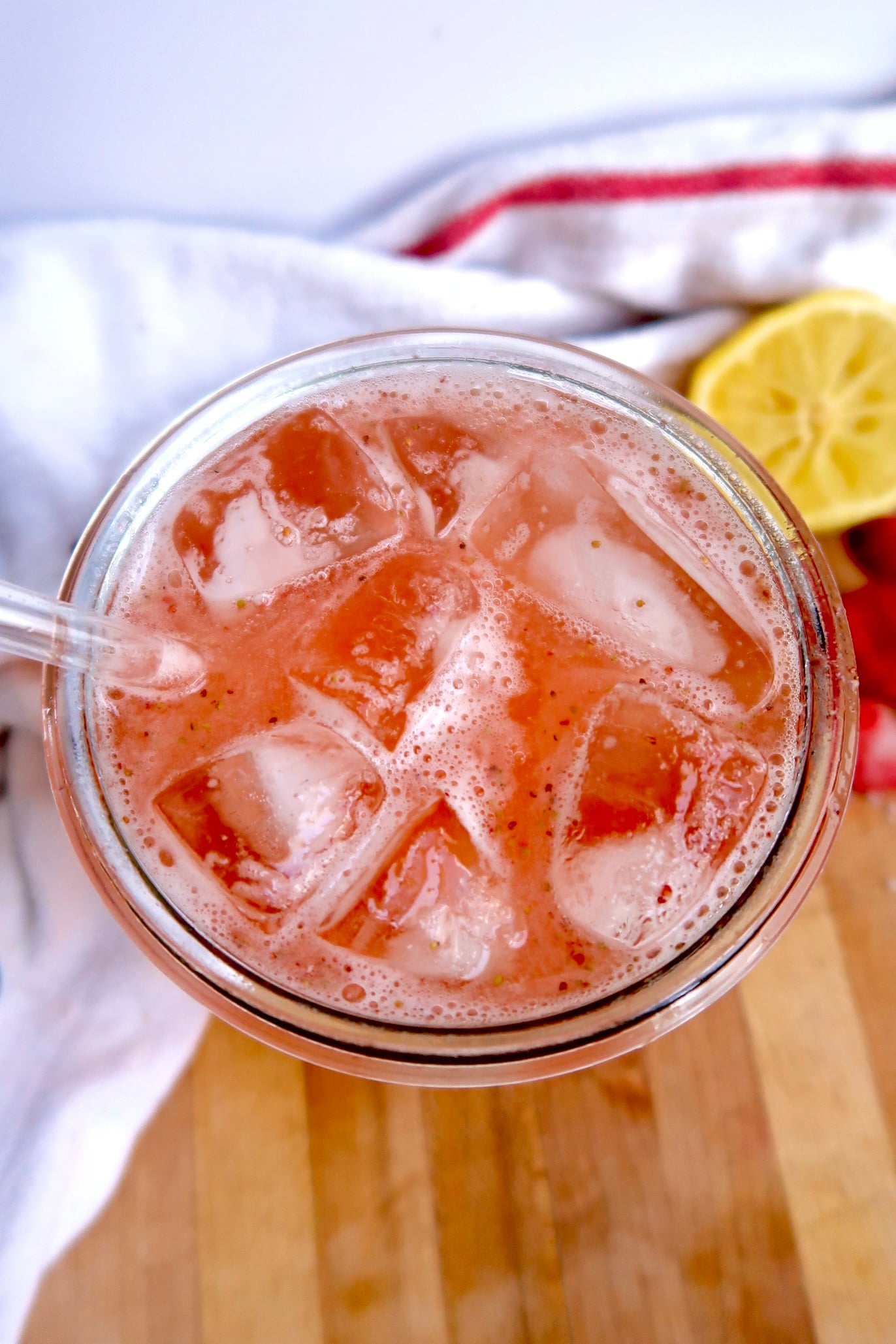 A glass of iced pink lemonade with a clear straw, viewed from above. The drink contains ice cubes and is set on a wooden surface with a white towel and a cut lemon in the background.