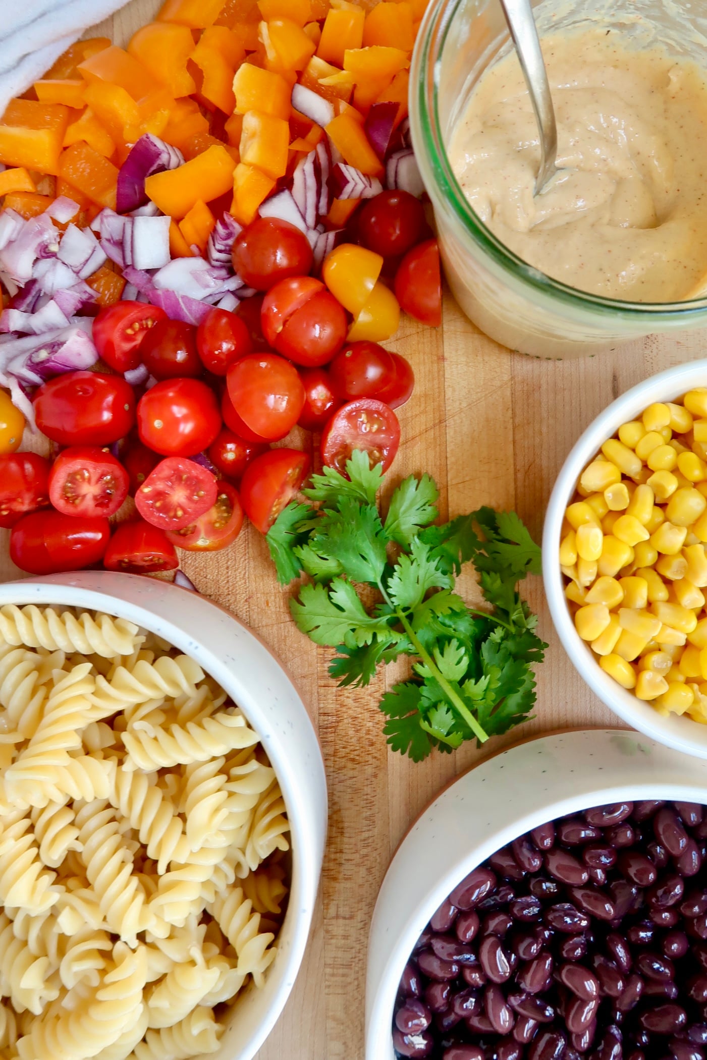 Cutting board with sliced cherry tomatoes, rotini pasta, creamy chipotle sauce, corn, cilantro and black beans.