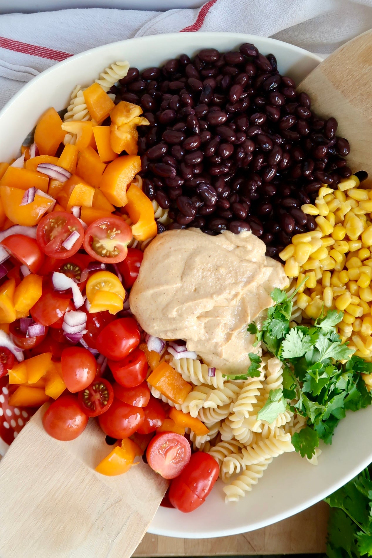 Large bowl with blac beans, corn, cilantro, rotini pasta, chopped bell peppers and cherry tomatoes with a creamy sauce in the center and tongs to the side.
