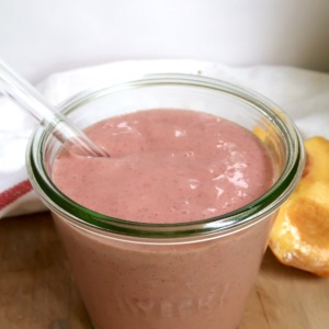 Pink smoothie in a jar with a glass straw and a peach to the side.