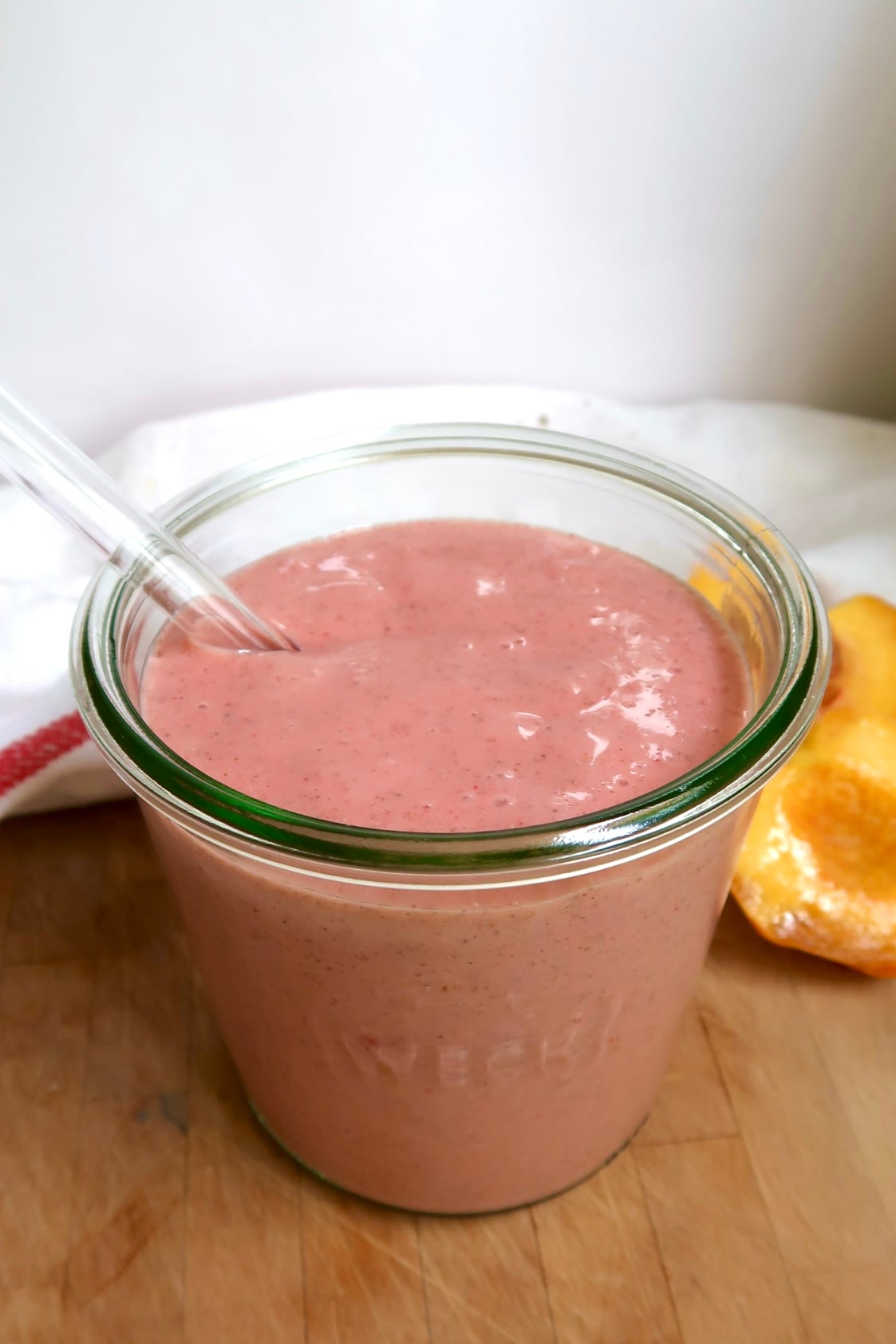 Pink smoothie in a jar with a glass straw and a peach to the side.