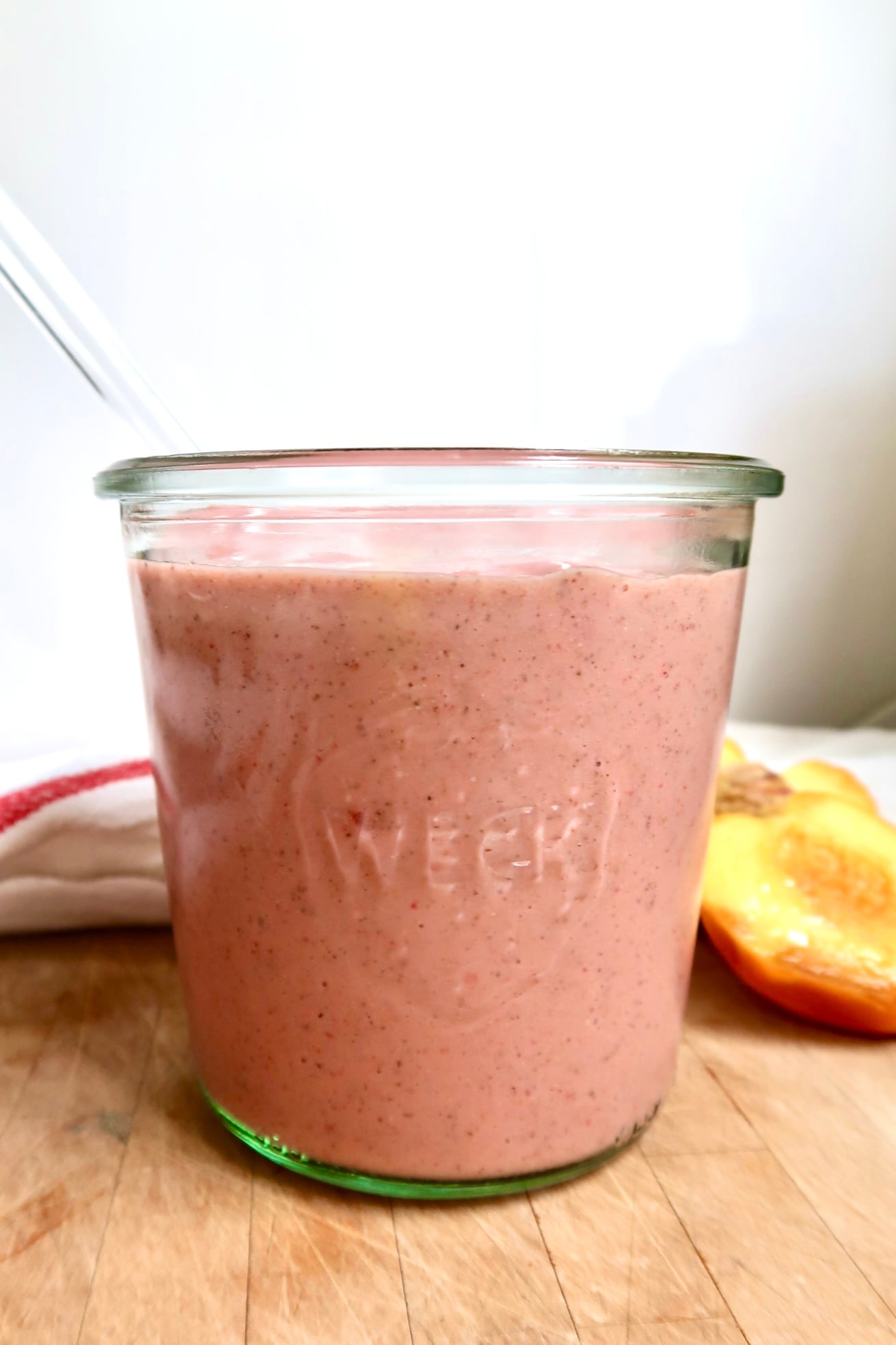 Pink smoothie in a jar with a glass straw in it and peaches to the side.
