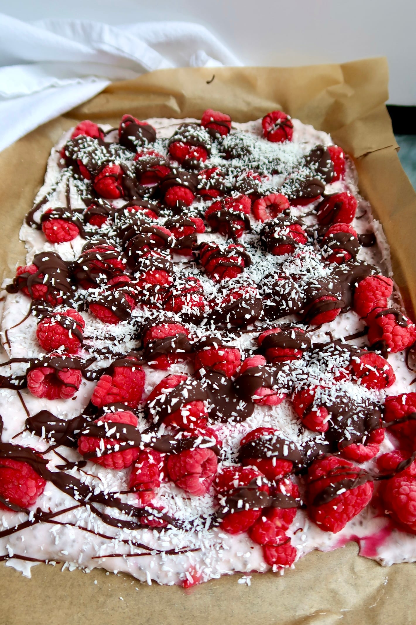 Side view of parchment paper with yogurt spread on top, raspberries, chocolate drizzled and coconut sprinkled on top.