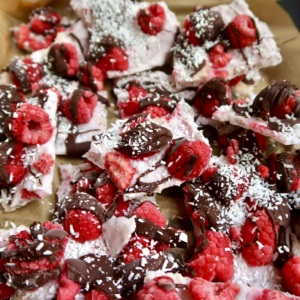 Parchment paper with yogurt spread on top, raspberries, chocolate drizzled and coconut sprinkled on top broken into pieces.