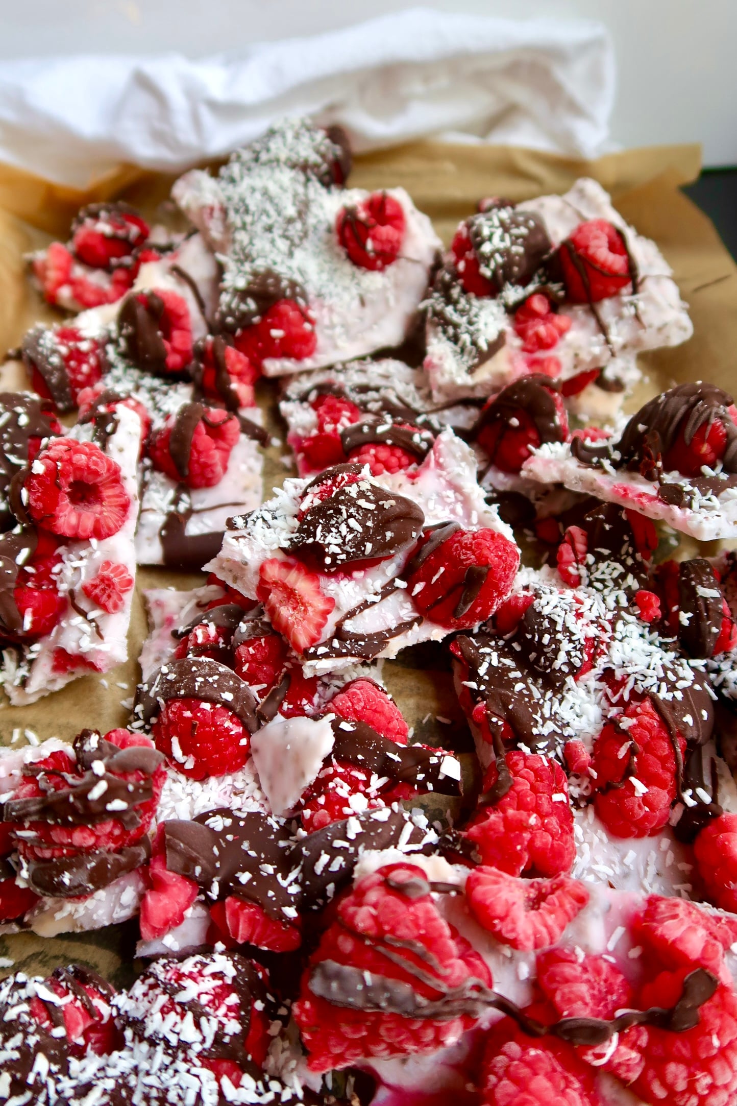 Parchment paper with yogurt spread on top, raspberries, chocolate drizzled and coconut sprinkled on top broken into pieces.
