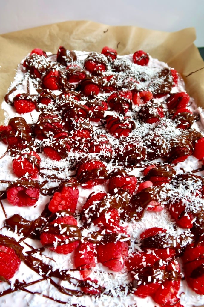 Side view of parchment paper with yogurt spread on top, raspberries, chocolate drizzled and coconut sprinkled on top.